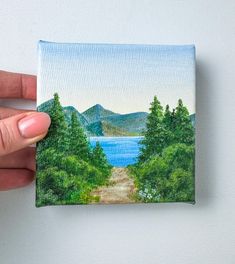 a hand holding up a small painting of a lake and mountains in the distance with trees on both sides