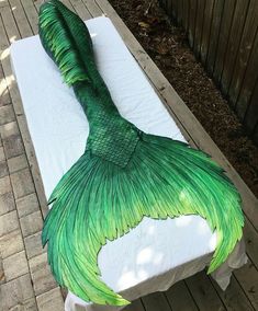 a large green mermaid tail laying on top of a white tablecloth covered bench in front of a wooden fence