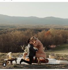 two people sitting on a blanket in the middle of a field with mountains in the background