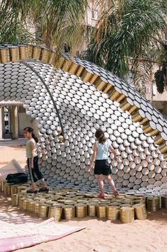 two people standing in front of a circular structure made out of beer cans on the beach