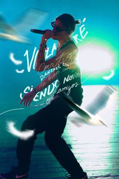 a man holding a microphone while standing on top of a wooden floor in front of a blue and green background