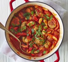 a red pot filled with meat and vegetables on top of a white table next to a wooden spoon