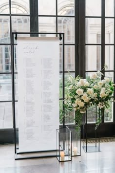 a wedding seating chart with flowers and candles in front of large windows at the reception