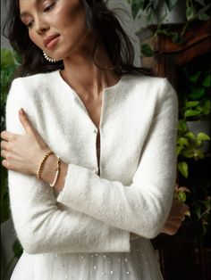 a woman with her arms crossed wearing a white cardigan sweater and gold bracelets