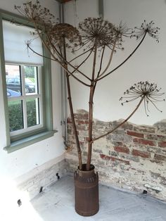 a tall plant in a brown vase next to a brick wall with a small window