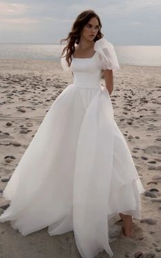 a woman standing on top of a sandy beach wearing a white dress and holding her hand in her pocket