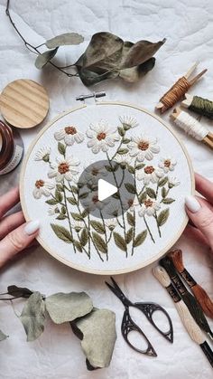 two hands are holding a embroidery project with flowers and scissors on the table next to it