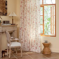 a room with a desk, chair and window covered in floral curtaining next to a painting