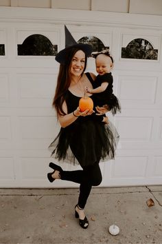 a woman holding a baby wearing a witch costume and holding an orange in front of a garage door