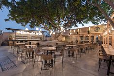 an outdoor restaurant with tables and stools under a large shade tree in the evening