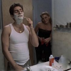 a man shaving his face in the bathroom while another woman looks at him through the mirror