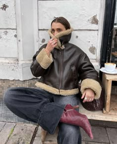 a woman is sitting on a bench with her hand in her mouth while wearing a jacket and boots