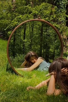 two women laying on the grass in front of a circular mirror with trees behind them