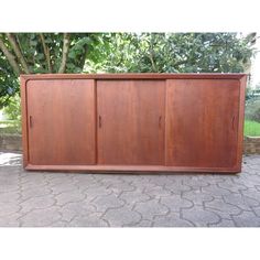 a large wooden cabinet sitting on top of a stone floor next to a green tree