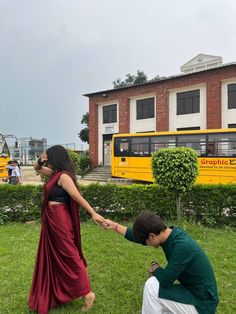 a man kneeling down next to a woman on top of a lush green field in front of a yellow school bus