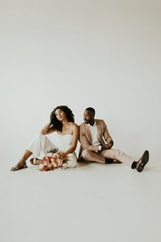 a man and woman sitting next to each other in front of a white wall with flowers on the floor