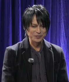 a man with black hair is sitting in front of a blue curtain