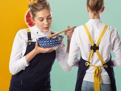 two women wearing aprons and holding chopsticks in front of their faces, one eating from a bowl