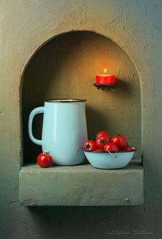 a bowl of tomatoes next to a candle on a shelf with a white mug in front of it