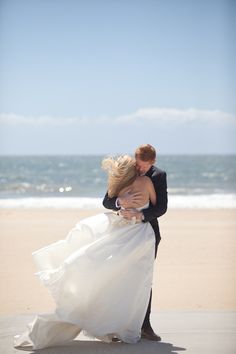 a bride and groom hug on the beach in front of the ocean while holding each other