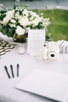 a white table topped with a camera next to a vase filled with flowers and writing utensils