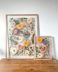 two framed pictures sitting on top of a wooden table next to each other with flowers painted on them