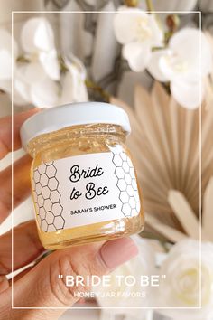 a hand holding a jar of bride to be honey with flowers in the back ground