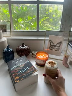 a person holding a drink in front of a book and candle on a table next to a window