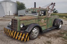 an old green tow truck parked in front of a grain silo on a gravel lot