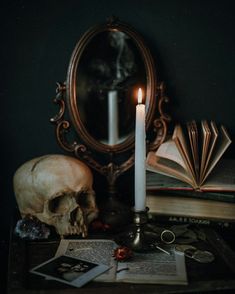 a candle is lit next to an open book and a skull on a table in front of a mirror