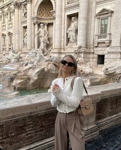 a woman standing in front of a fountain with her hand on her hip and wearing sunglasses