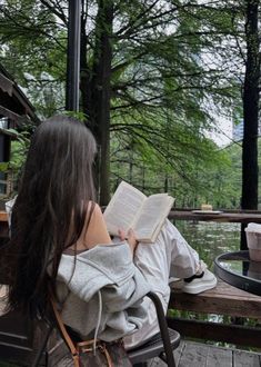 a woman reading a book while sitting on a bench in front of a lake and trees