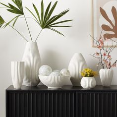 several white vases with plants in them on a black cabinet next to a painting