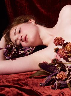 a naked woman laying on top of a bed next to purple and red flowers with her eyes closed