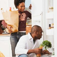 a man and woman in the kitchen looking at each other's vegetables while they are holding paper bags