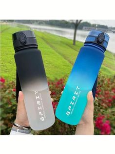 two water bottles are held up in front of some bushes and flowers, one is blue and the other is black