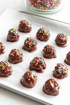 chocolate covered donuts with sprinkles are on a baking sheet next to a jar of sprinkles