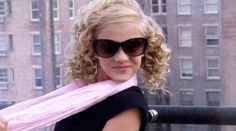 a woman with curly hair wearing sunglasses and a pink scarf around her neck smiles at the camera