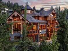 a large wooden house surrounded by trees in the woods at dusk with lights on it's windows