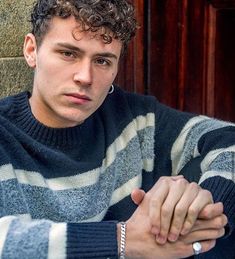 a young man with curly hair wearing a striped sweater sitting in front of a brick wall