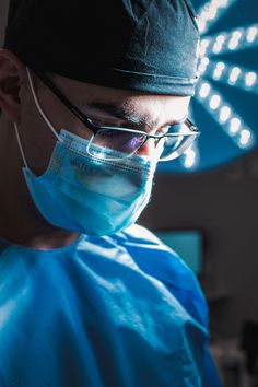 a surgeon in scrubs and glasses looking down at something on the table behind him