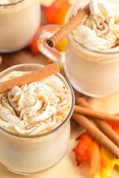 two mugs filled with whipped cream and cinnamon on a wooden table next to autumn leaves