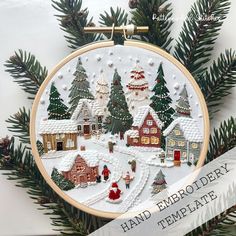 a hand embroidered christmas ornament hanging on a tree branch with snow covered houses and evergreens