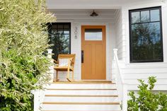 a white house with a wooden door and steps leading up to the front door is surrounded by greenery