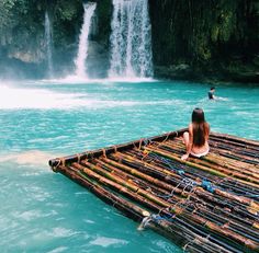 a woman sitting on top of a bamboo raft in front of a waterfall with people swimming