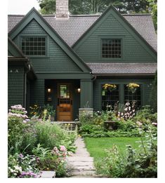 a green house with lots of flowers in the front yard and bushes on either side