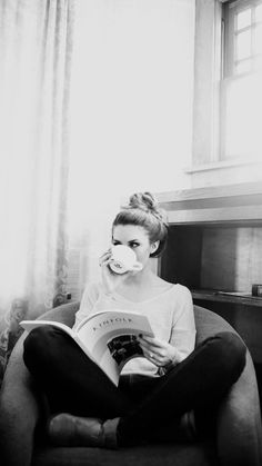 a woman sitting in a chair drinking from a cup while reading a book with her eyes closed