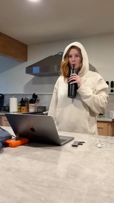 a woman in a hoodie drinking from a cup while using a laptop