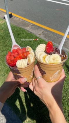 two people are holding up ice cream with strawberries and bananas in their cups on the grass