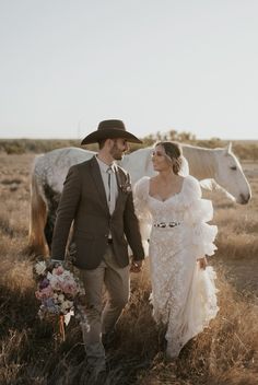 a bride and groom are walking in the field with their horse behind them, holding hands